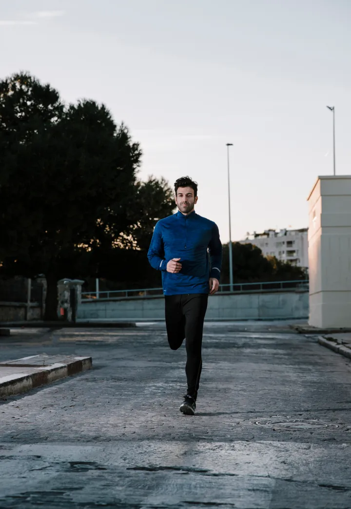 Homem corre em rua pavimentada ao ar livre vestindo blusa azul e calça preta.
